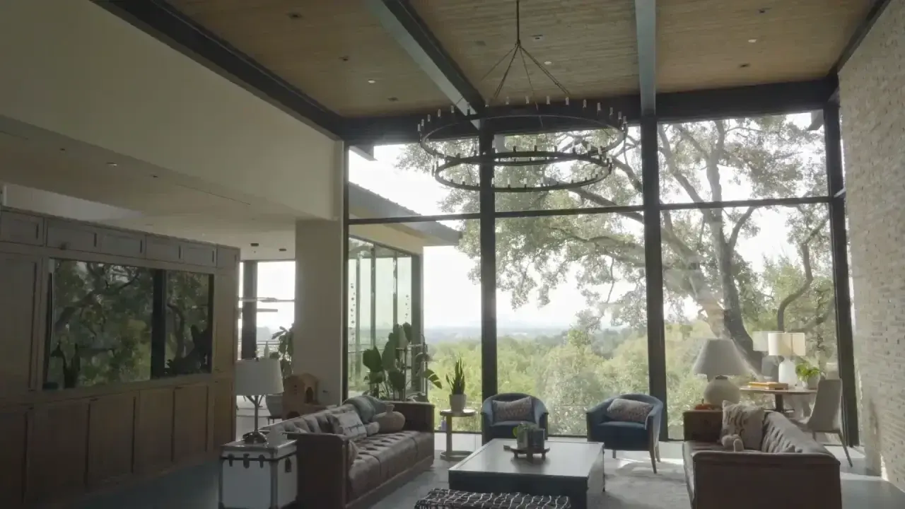 Glass-walled living room with a ring chandelier and treetop views. Lighting and motorized shade installation in Bee Cave, TX by AV Connect.