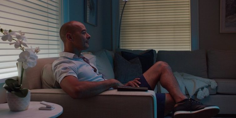 man lounging on a couch next to a lutron pico remote on a coffee table. automated blinds are lowered in the background.