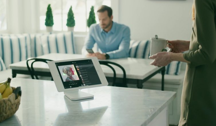 modern kitchen with a tablet on the countertop displaying a music playlist, a man in the background, and a woman holding a cup of tea.